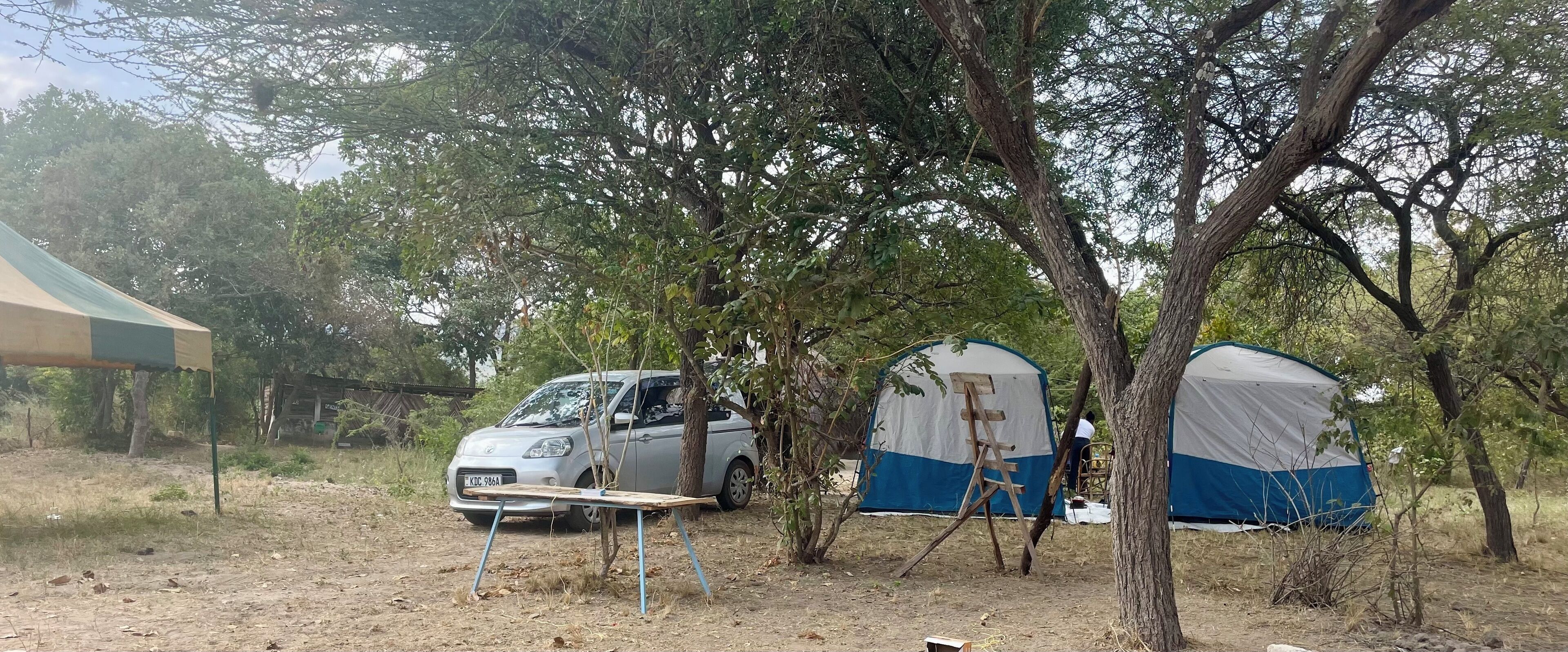 Un campamento con dos tiendas de campaña, un coche y una mesa debajo de un árbol.