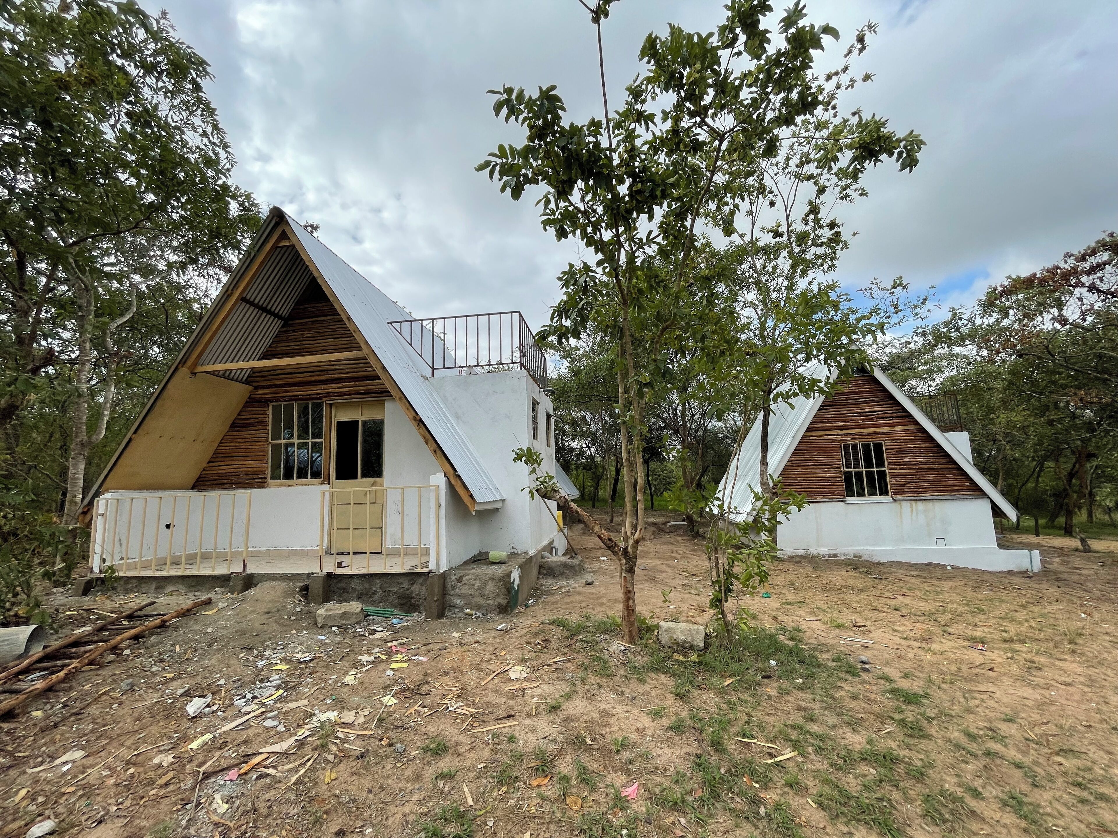 Deux chalets en forme de « A », avec des façades en bois et des toits en métal, entourés d’arbres et situés sur un terrain en terre battue.