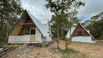 Deux chalets en forme de « A », avec des façades en bois et des toits en métal, entourés d’arbres et situés sur un terrain en terre battue.