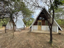 Two A-frame cabins with white railings, surrounded by trees and a dirt ground.