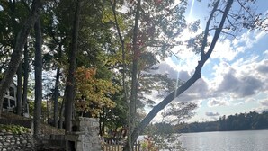 Une terrasse en bois avec des chaises en métal, donnant sur un lac, entourée d’arbres et d’un mur de pierre.