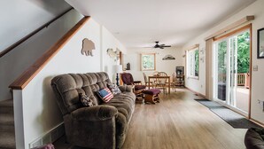 Een ruime woonkamer met een houten trap, een grote bruine bank, een eettafel met stoelen en uitzicht door de glazen deuren.