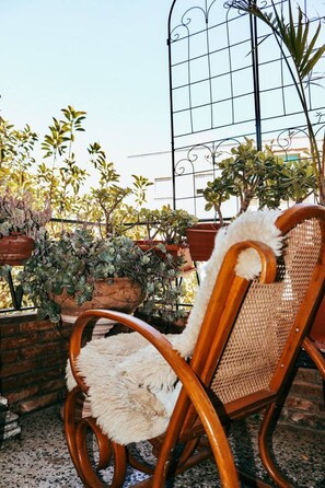 A wicker chair with a fur seat and backrest, placed on a balcony with potted plants and a metal railing.