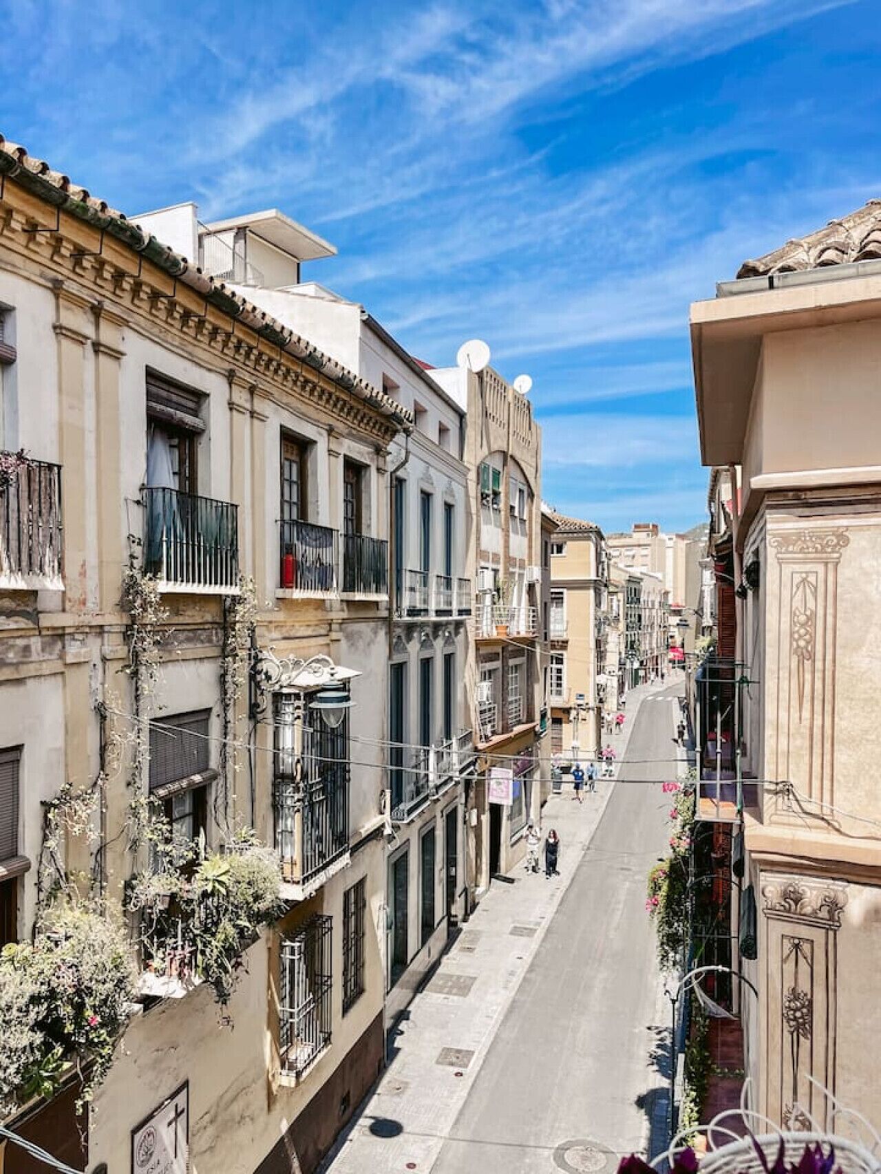 A narrow street lined with historic buildings, featuring balconies with plants and satellite dishes, under a clear blue sky.