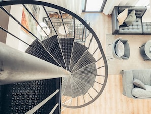 A spiral staircase with a black metal railing and a wooden floor. There is a gray sofa and a round table with a cushion in the room below. - Panoramic Rooftop Duplex Retreat | Free Parking (Málaga)