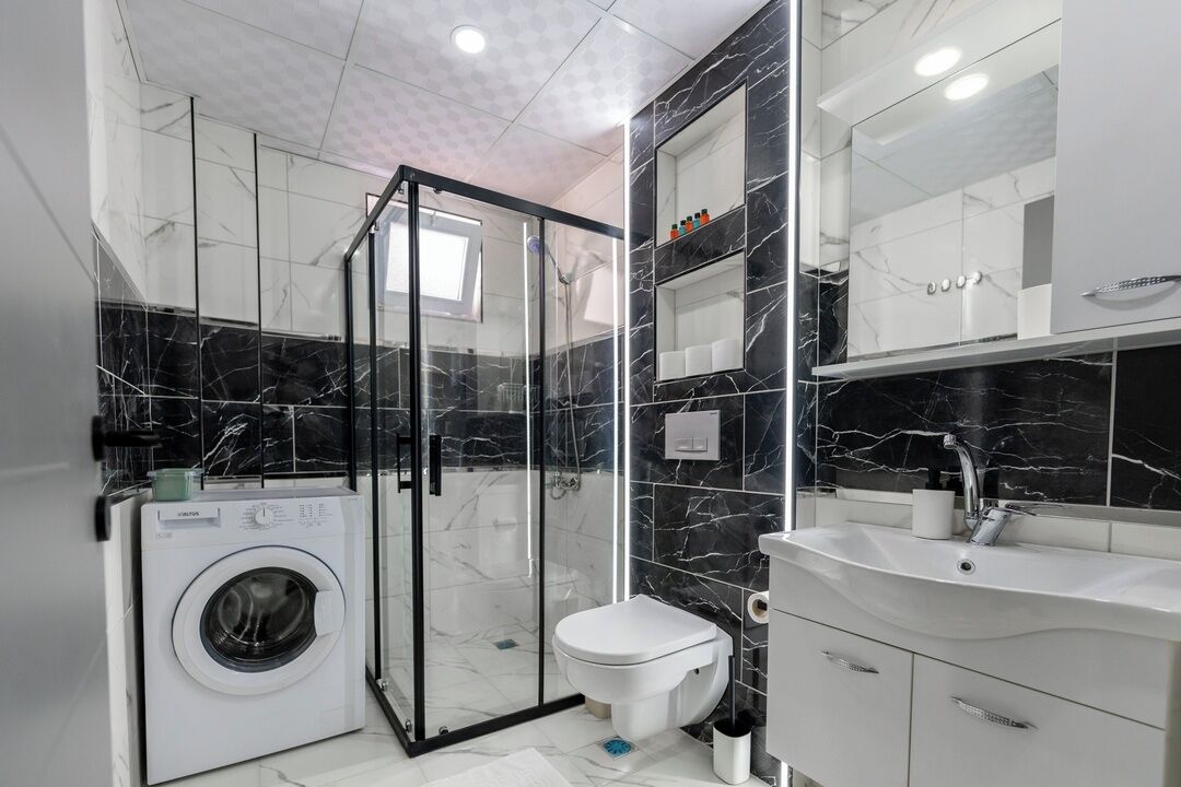 Une salle de bain moderne dotée d’une douche à parois de verre, d’un lave-linge encastré, de toilettes et d’un meuble-lavabo avec un évier et un miroir.