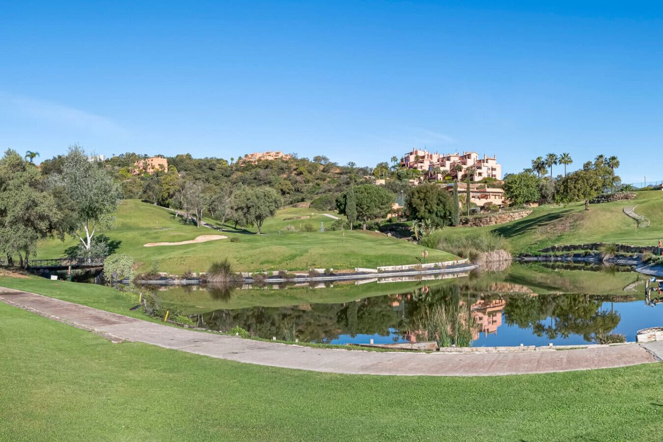A golf course with a pond, a bridge, and buildings in the background.
