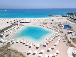 Eine Luftaufnahme eines Strandresorts mit einem zentralen Pool, Liegestühlen und einem Pier, der ins Meer ragt.