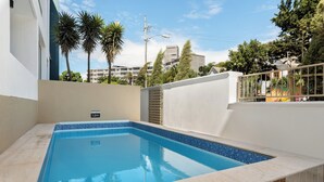 Una piscina rectangular con borde de azulejos, rodeada por un muro blanco y una terraza de baldosas color beige.
