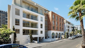 Un edificio moderno de varios pisos con amplios ventanales y balcones, un estacionamiento con coches y palmeras.