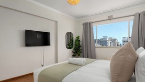 A modern hotel room with a large bed, a flat-screen TV mounted on the wall, a window with curtains, and a city view. - Heart of Seapoint Hideaway (Cape Town)