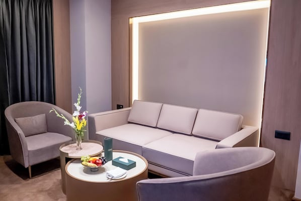 A modern hotel room with a beige sofa, two armchairs, a round coffee table with fruits and a vase of flowers, and a large illuminated wall panel. - ASTER GRAND BY ZENQ SAKET (New Delhi)