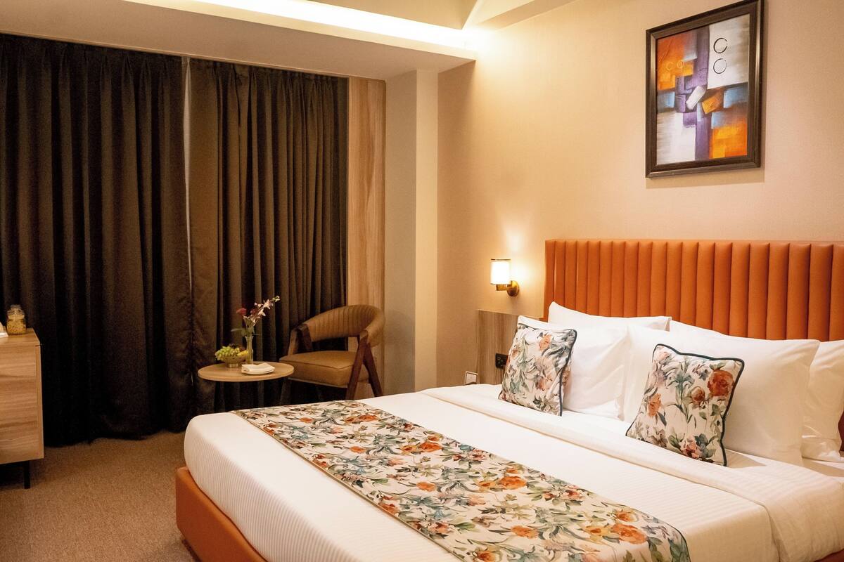 A hotel room with a large bed, a chair, a small table with a vase of flowers, and a painting on the wall.