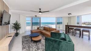 A modern living room with a large window overlooking a beach, a ceiling fan, and a dining area with a view.