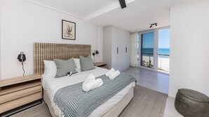 A modern bedroom with a large bed, a headboard, bedside lamps, a view of the beach, and a ceiling fan.