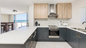 A modern kitchen with grey cabinets, a white countertop, and stainless steel appliances. There is a view of the ocean through a large window.