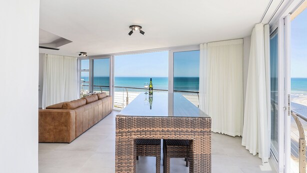 A modern living room with a large glass table, a brown leather sofa, and a view of the ocean.