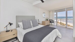 A modern bedroom with a large bed, a nightstand, a desk, and a view of the beach.