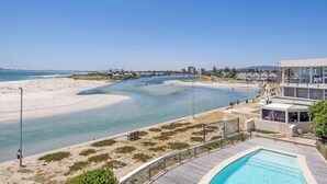 A coastal view with a beach, a swimming pool, and a modern building with large windows.