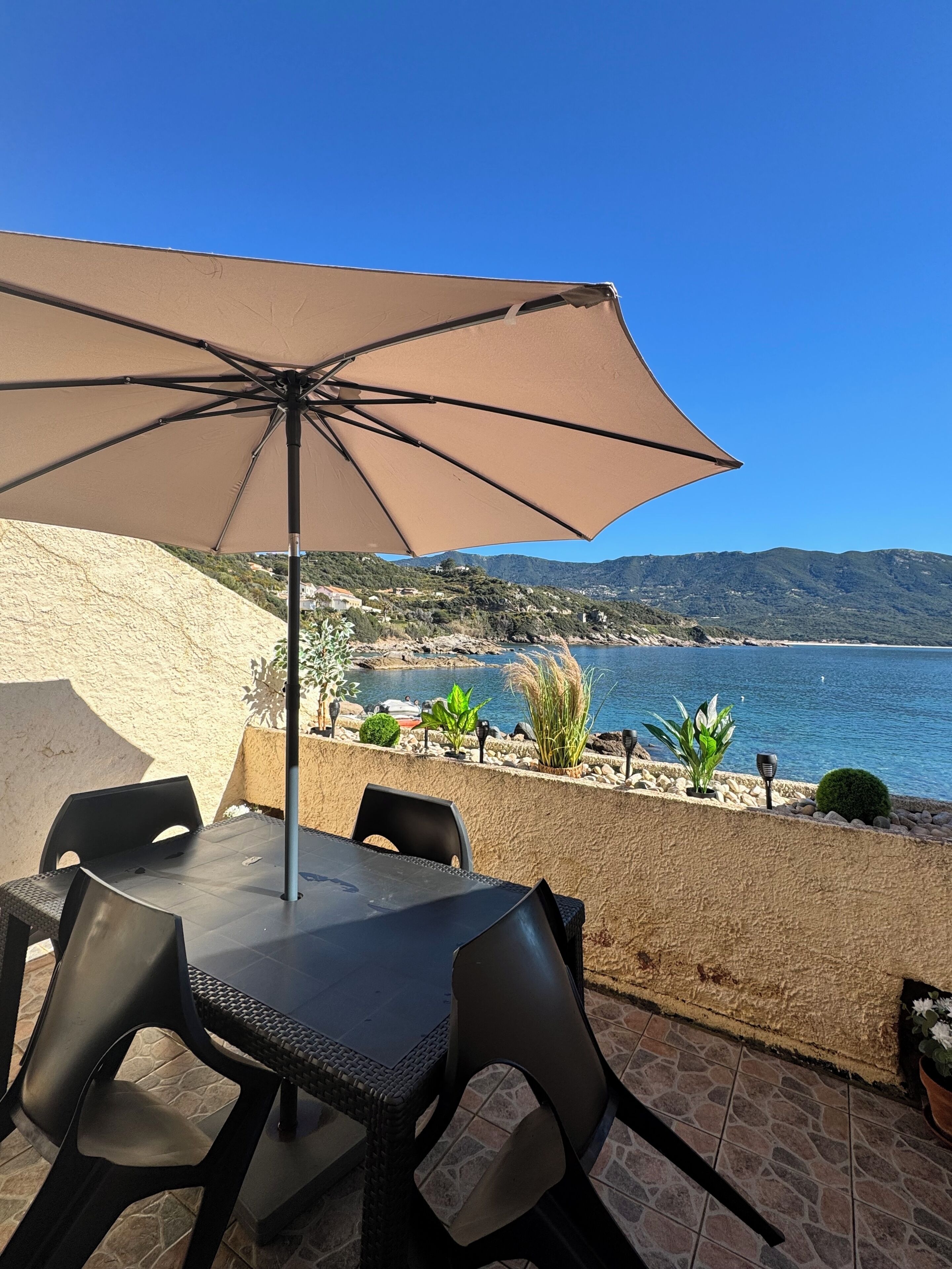 An outdoor patio with a table and chairs, a beige umbrella, and a view of the sea and mountains.