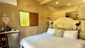 Ein Schlafzimmer mit einem großen Bett, einem Deckenventilator, einem kleinen Schreibtisch mit Kühlschrank, einem Nachttisch und einem Fenster mit Blick auf Grünflächen.