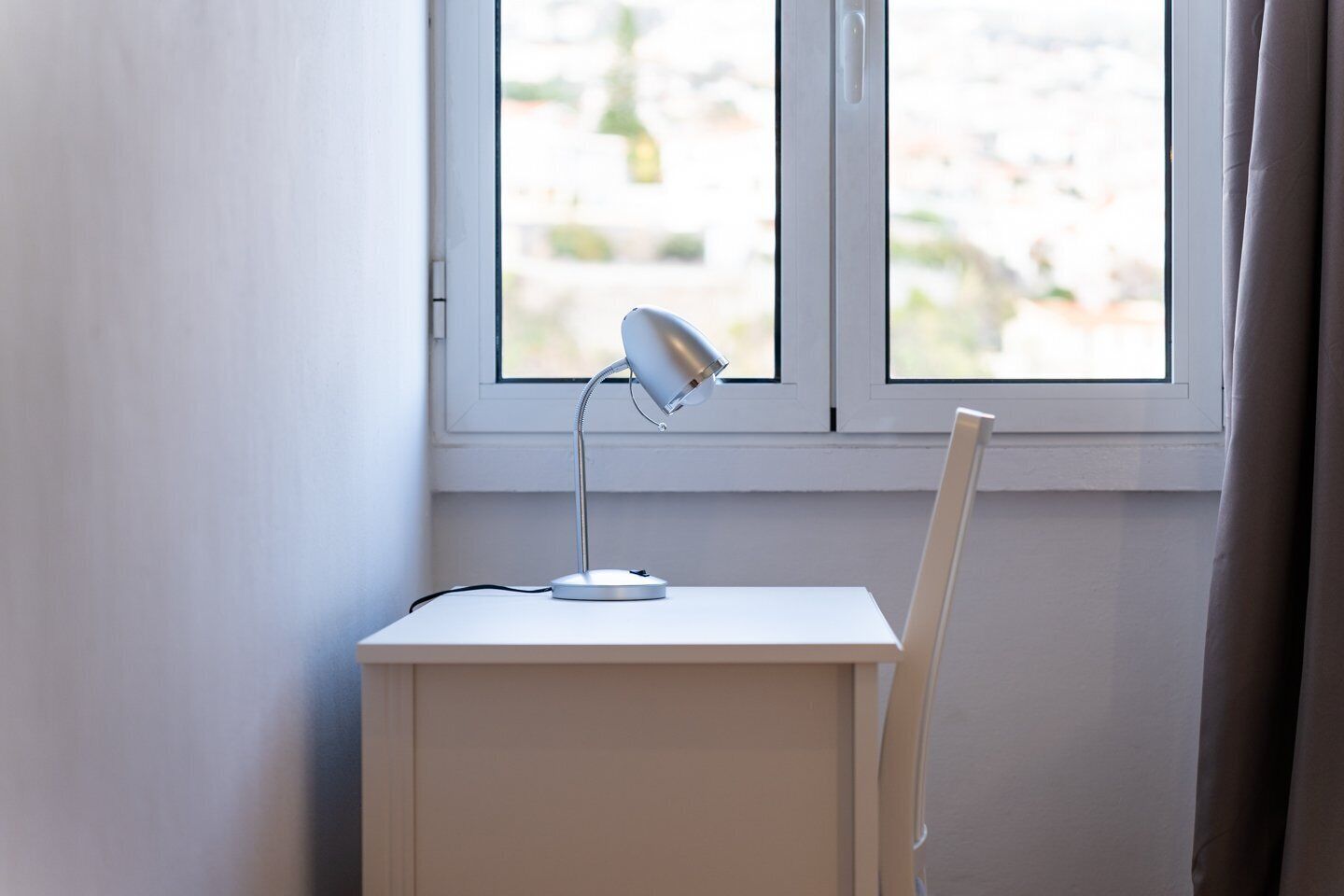 Uma secretária branca com uma lâmpada de mesa e uma cadeira em frente a uma janela com molduras brancas.