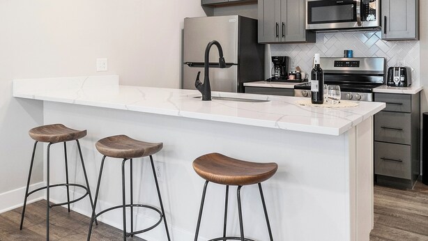 A modern kitchen with grey cabinets, a white countertop, and three stools.