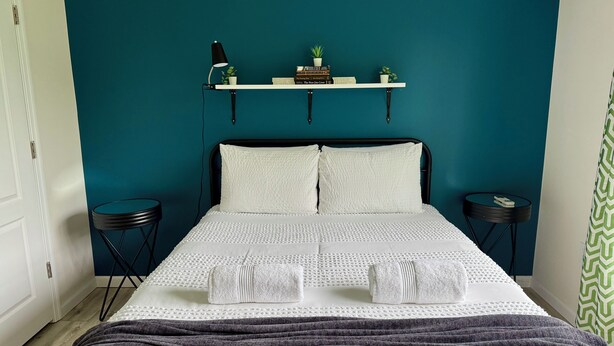 A neatly made bed with white linens and two pillows, a bedside table with a lamp, a small shelf with books and a plant, and a green patterned curtain.
