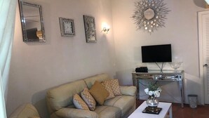 Una sala de estar con un sofá color beige, un televisor de pantalla plana, un espejo decorativo y una lámpara de pared.