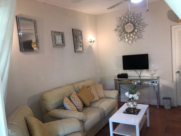 Una sala de estar con un sofá color beige, un televisor de pantalla plana, un espejo decorativo y una lámpara de pared.