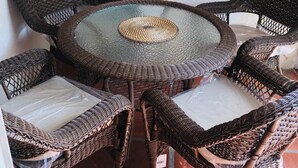 Una mesa redonda de mimbre con un tapete tejido en el centro, rodeada por cuatro sillas de mimbre con cojines blancos. Un espejo decorativo con forma de sol cuelga en la pared sobre la mesa.