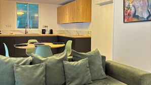A modern living room with a green sofa, a dining area with a table and chairs, a kitchen with wooden cabinets, and a large window.