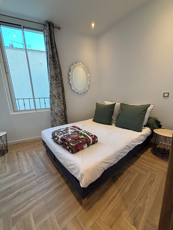 A bedroom with a large bed, a window with curtains, a wall-mounted mirror, and a small round table.
