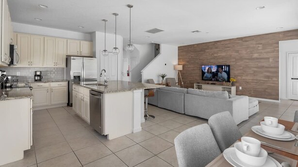 A modern kitchen with a central island, a dining area with a table set for two, and a living room with a sofa and a TV. - Amazing House at Bella Vida 5BR (Kissimmee)
