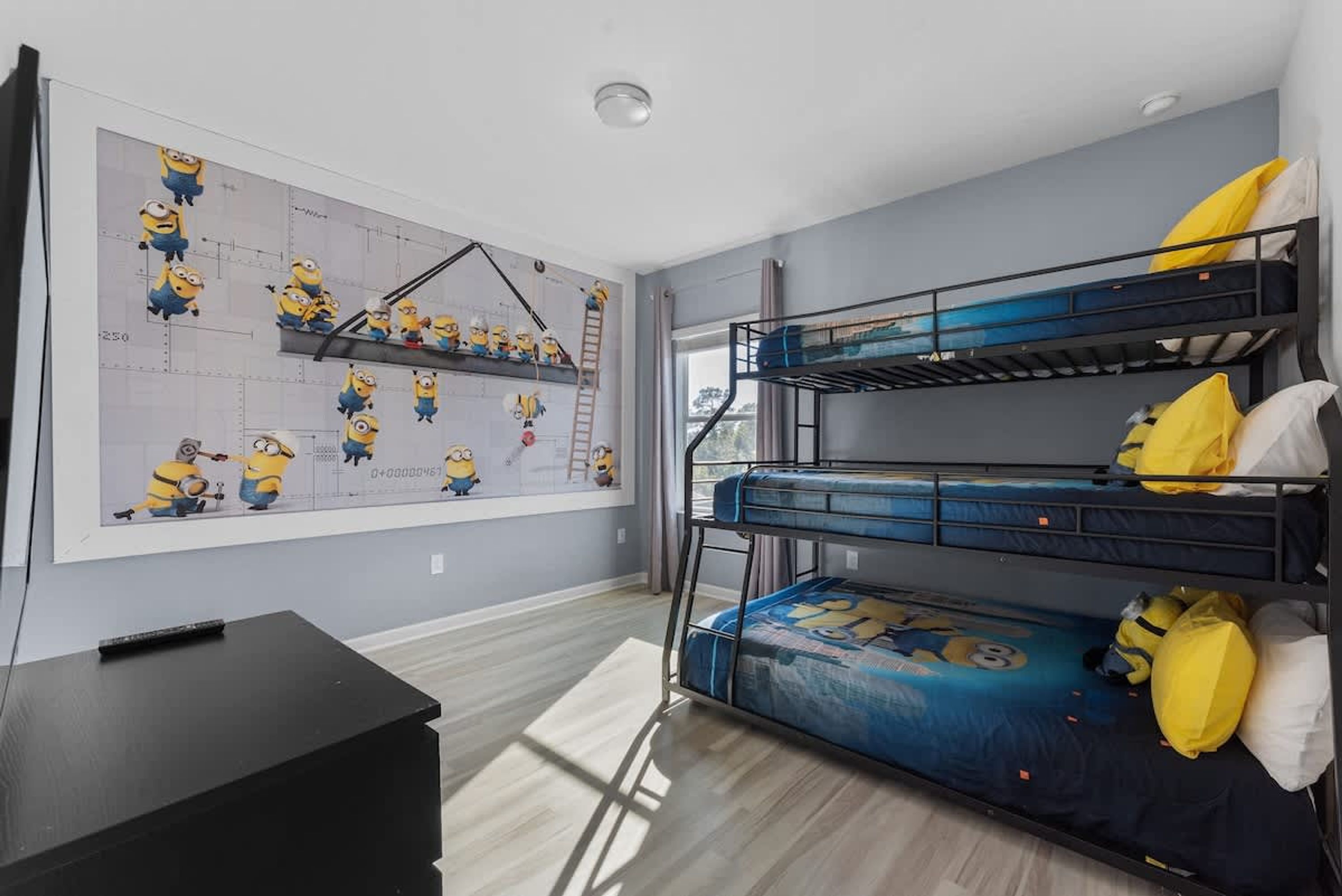 Une chambre avec un lit superposé, un mur décoré sur le thème des Minions, un bureau noir et une fenêtre avec des rideaux.