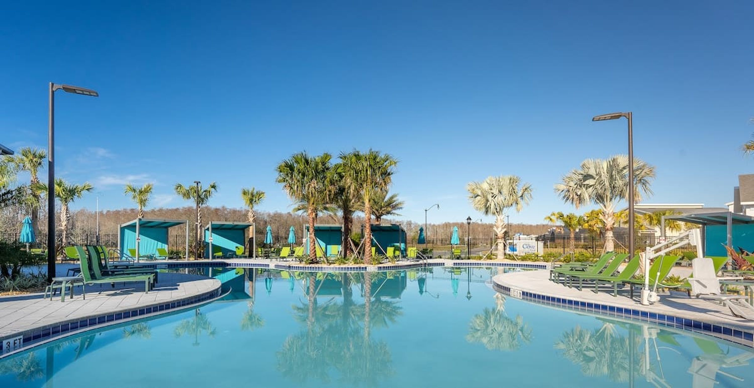 A clear blue pool surrounded by lounge chairs and umbrellas, with palm trees and a clear sky in the background.