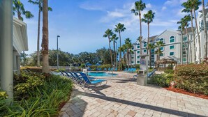 A hotel pool area with lounge chairs, palm trees, and a building with multiple windows. - 1BR Free Shuttle to Disney Parks (Orlando)