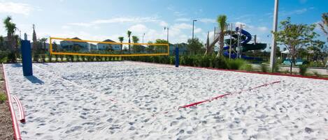 A sandy beach volleyball court with a net, surrounded by palm trees and a water slide in the background.