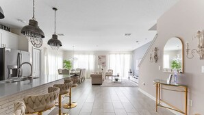 A modern kitchen with a central island, bar stools, pendant lighting, and a dining area with chairs and a sofa.