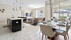 A modern kitchen with a central island, a dining area with a glass table, and a living space with a sofa and TV.