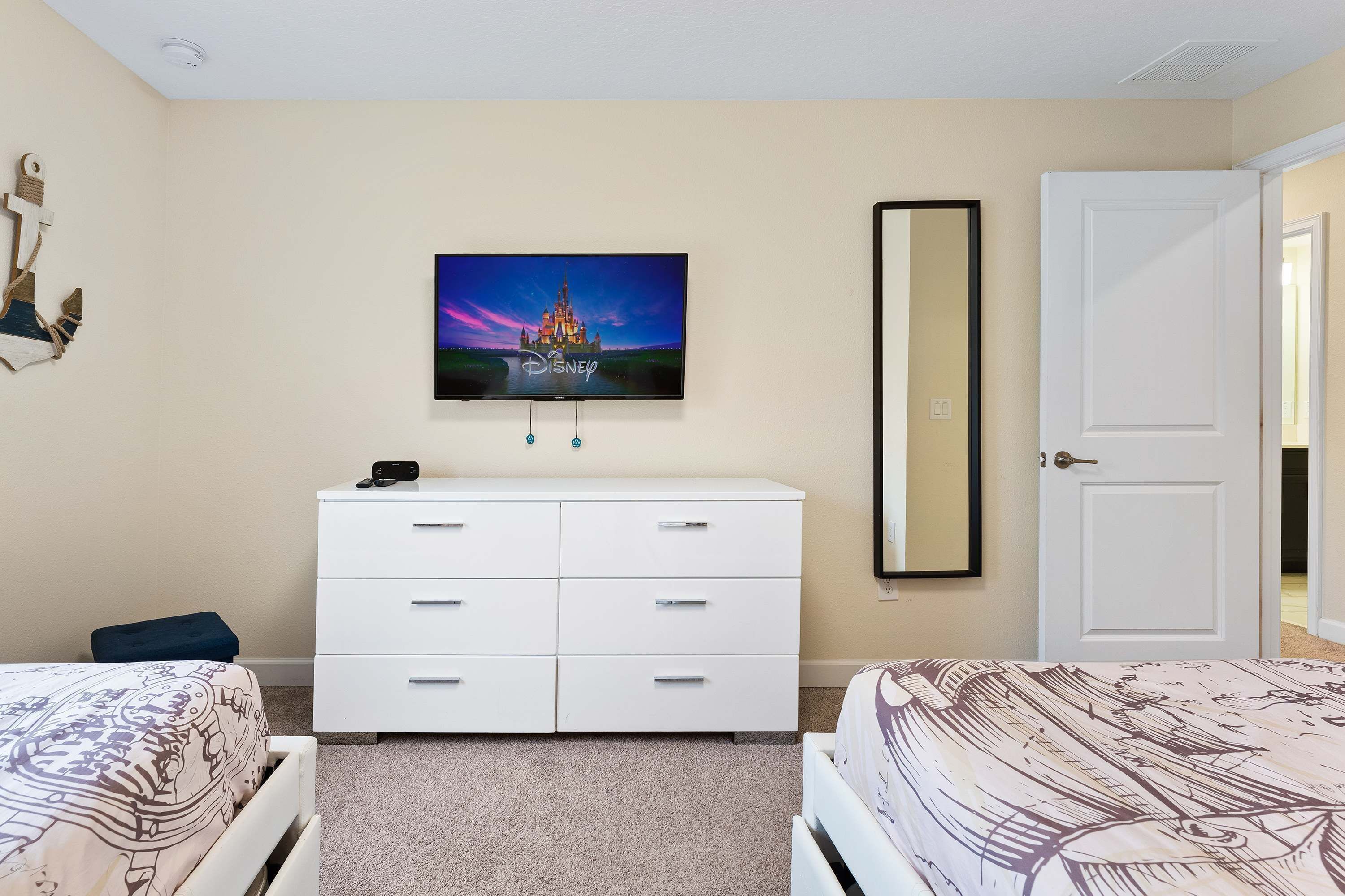 A bedroom with two beds, a white dresser, a TV mounted on the wall displaying a Disney castle, and a door to the right.