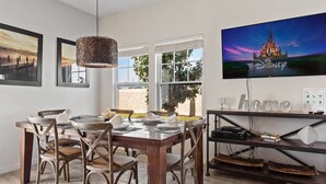 A dining area with a wooden table set for four, surrounded by wicker chairs. A television mounted on the wall displays a Disney castle. - Modern Decor at West Lucaya 4 BR Wl8989 (Kissimmee)