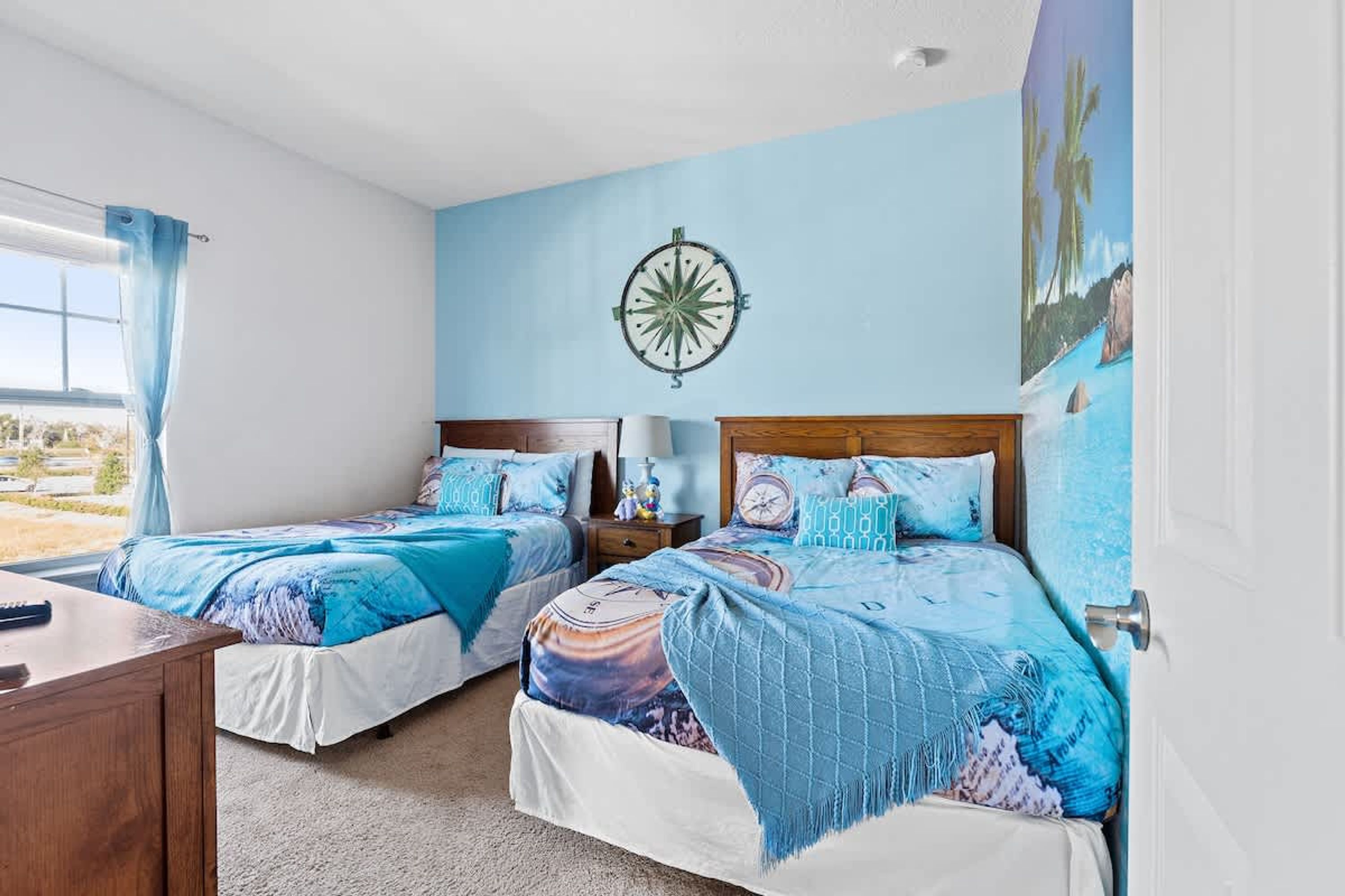 A bedroom with two beds, a wooden nightstand, a desk, a compass wall decoration, and a large window with curtains.