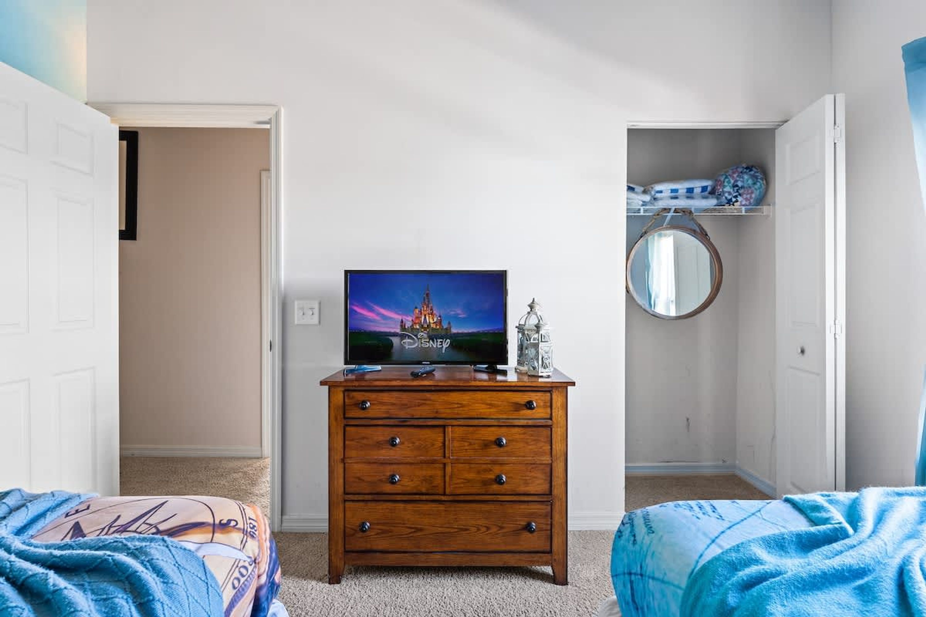 Une chambre avec une commode en bois, un téléviseur diffusant un film de Disney, un miroir rond et une couverture bleue sur le lit.