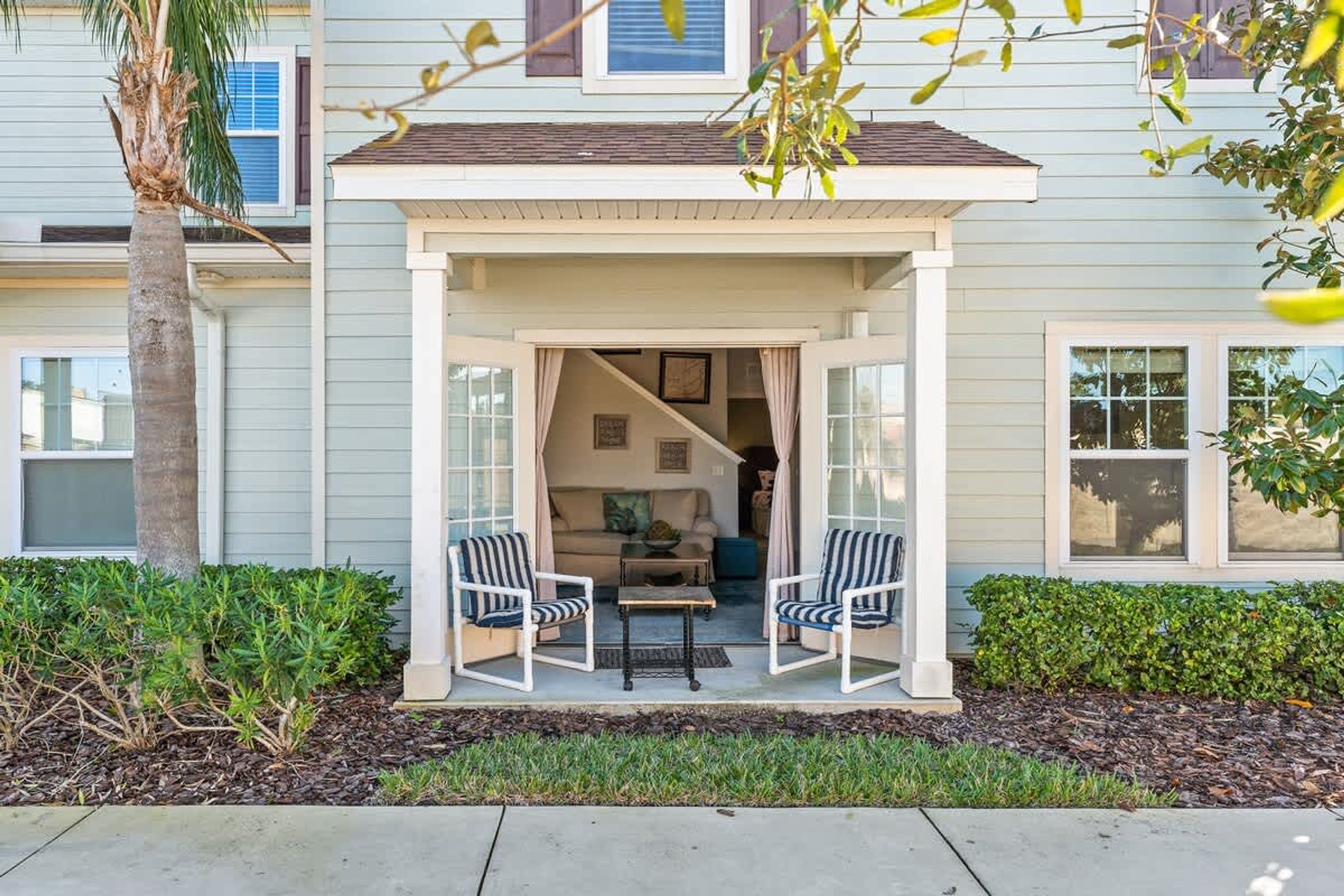A covered porch with two chairs and a table, a palm tree, and a house with a light blue exterior and white trim.