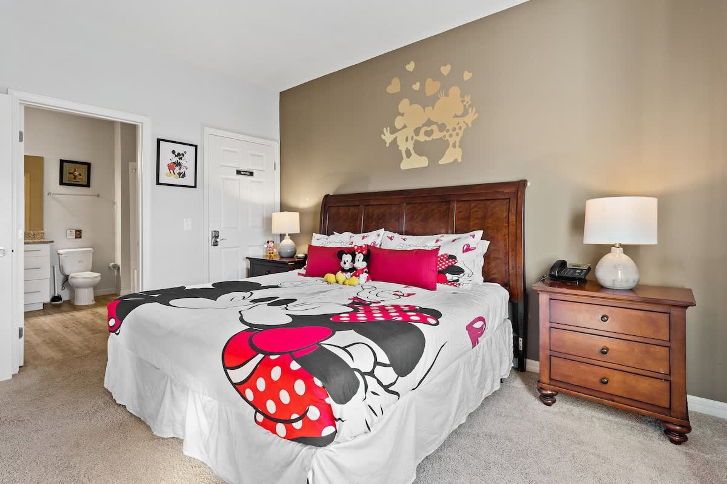 Une chambre à coucher avec un lit orné d’une couvre-lit aux motifs de Minnie Mouse, une table de chevet en bois avec une lampe, et un mur décoré d’un autocollant en forme de cœur.