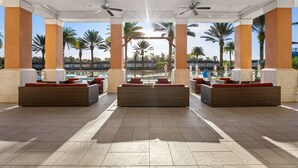 A covered outdoor lounge area with seating and ceiling fans, palm trees in the background, and a view of a pool and buildings.