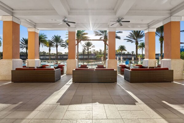 A covered outdoor lounge area with seating and ceiling fans, palm trees in the background, and a view of a pool and buildings.
