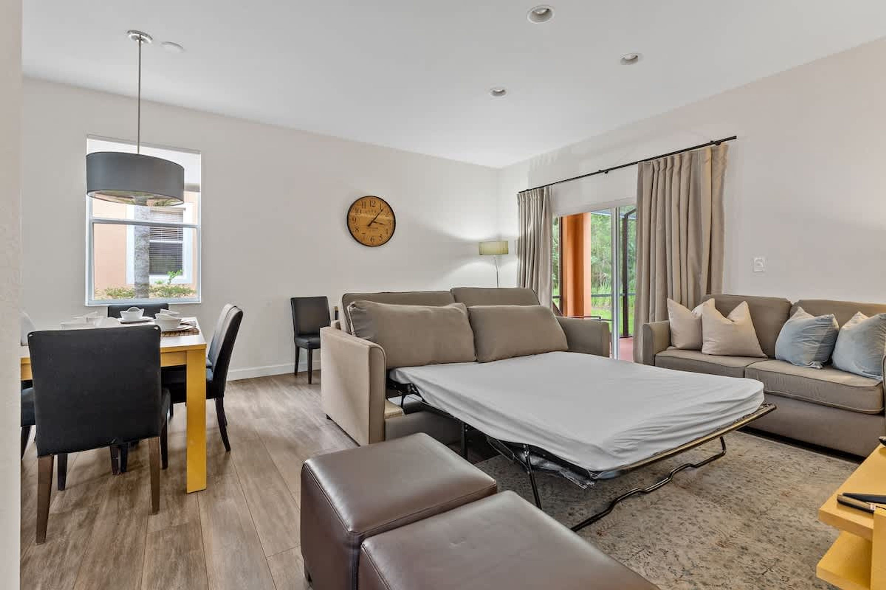 A modern living room with a sofa, a dining table with chairs, a bed, and a large window with curtains.