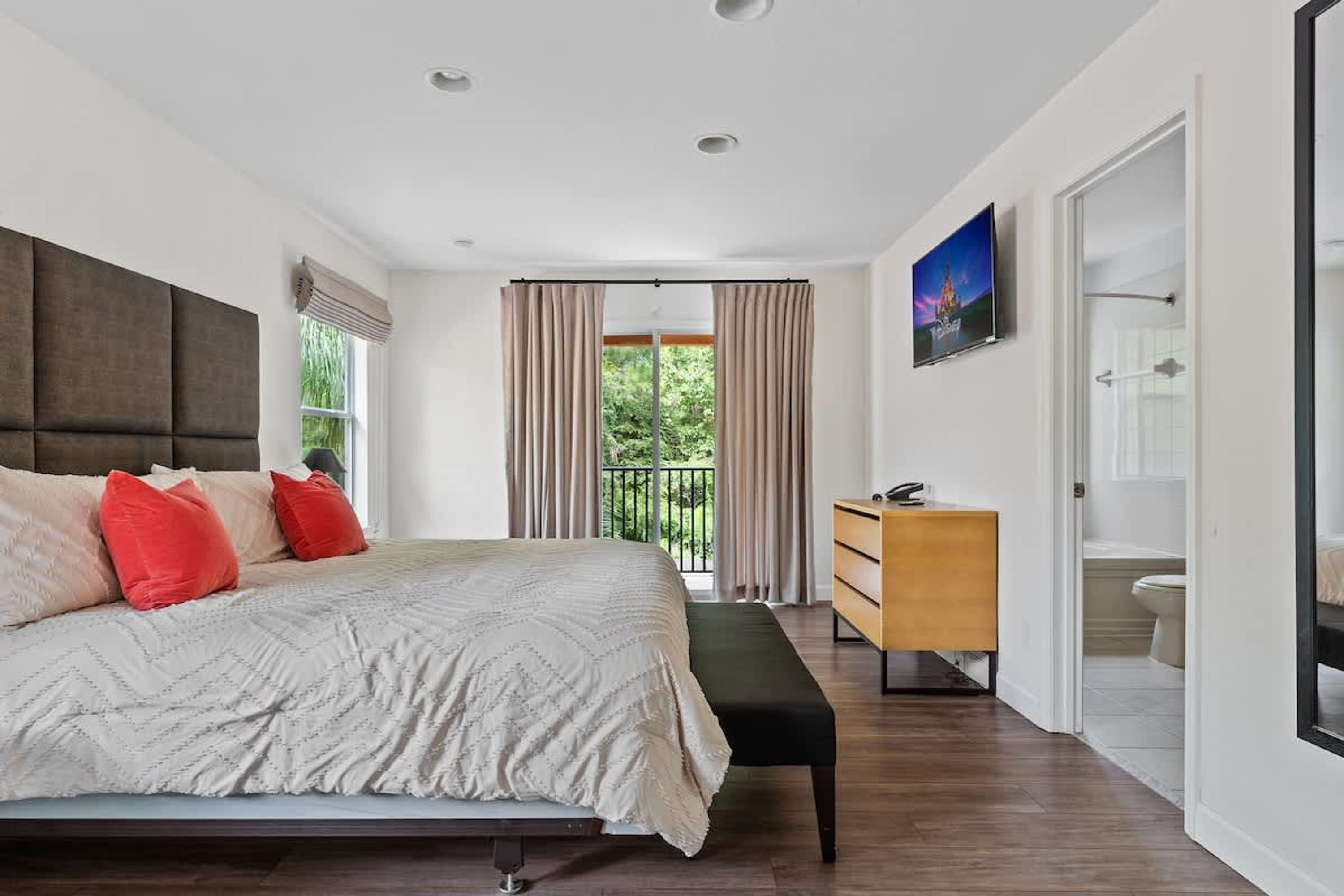 Une chambre moderne avec un grand lit, une commode en bois et une vue sur la verdure depuis un balcon.
